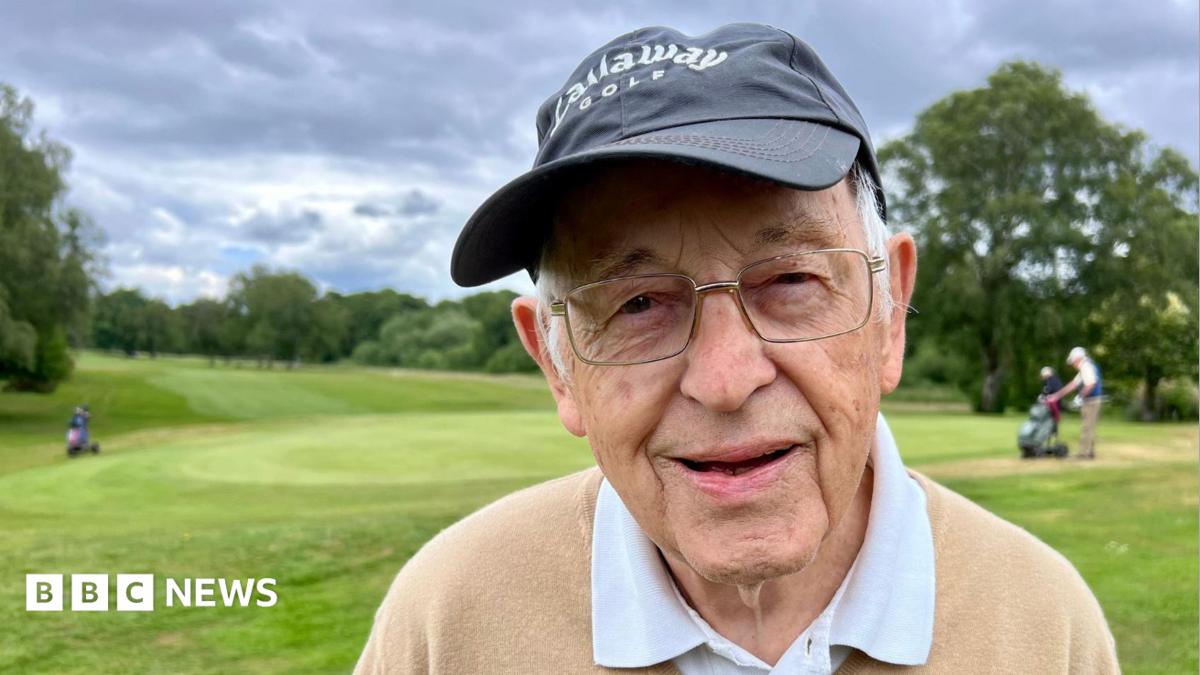 An elderly man with glasses and a black cap. He is looking into the camera. The picture is taken on a golf course.