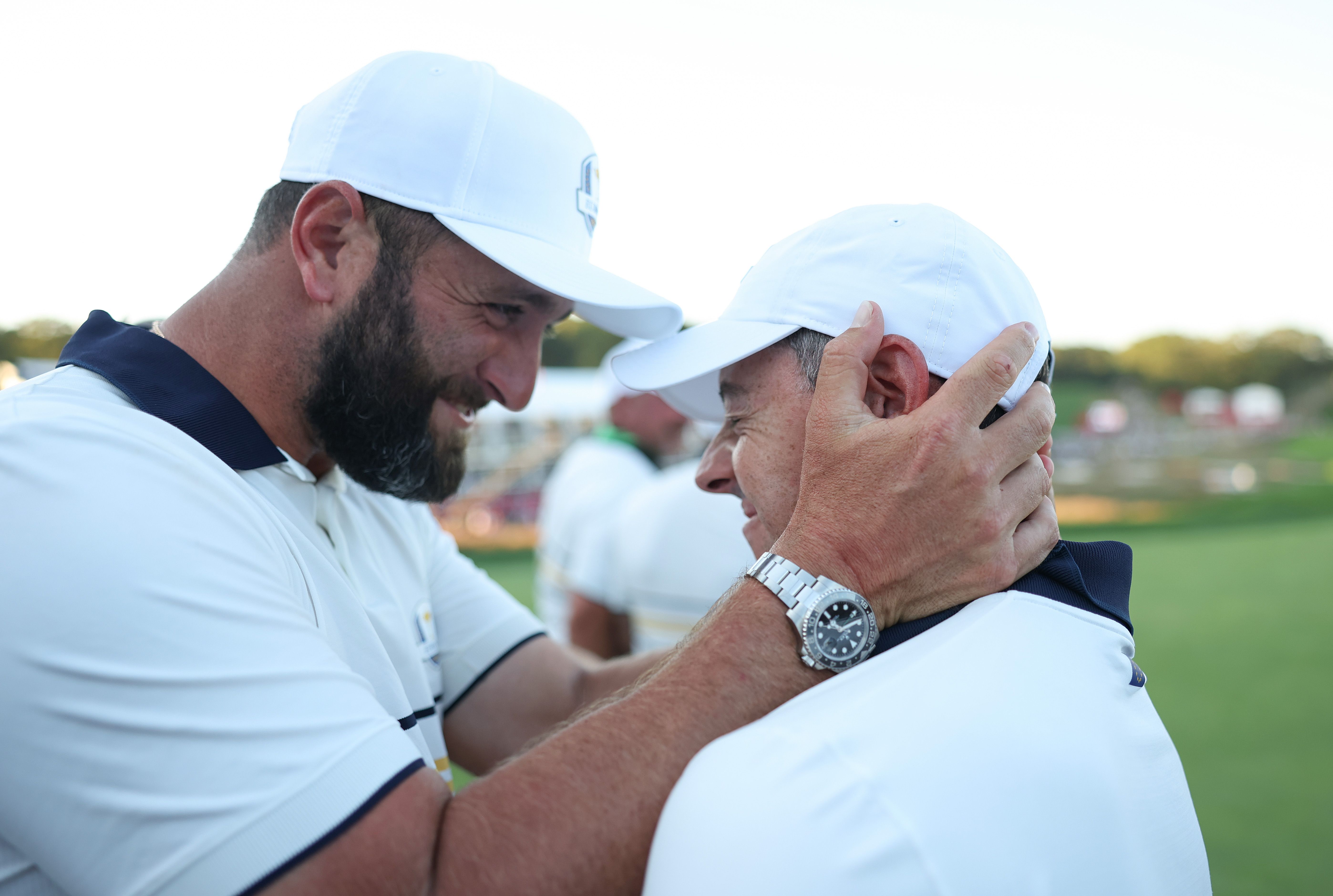 A smiling bearded man adjusts another man's white hat on a sunny golf course.