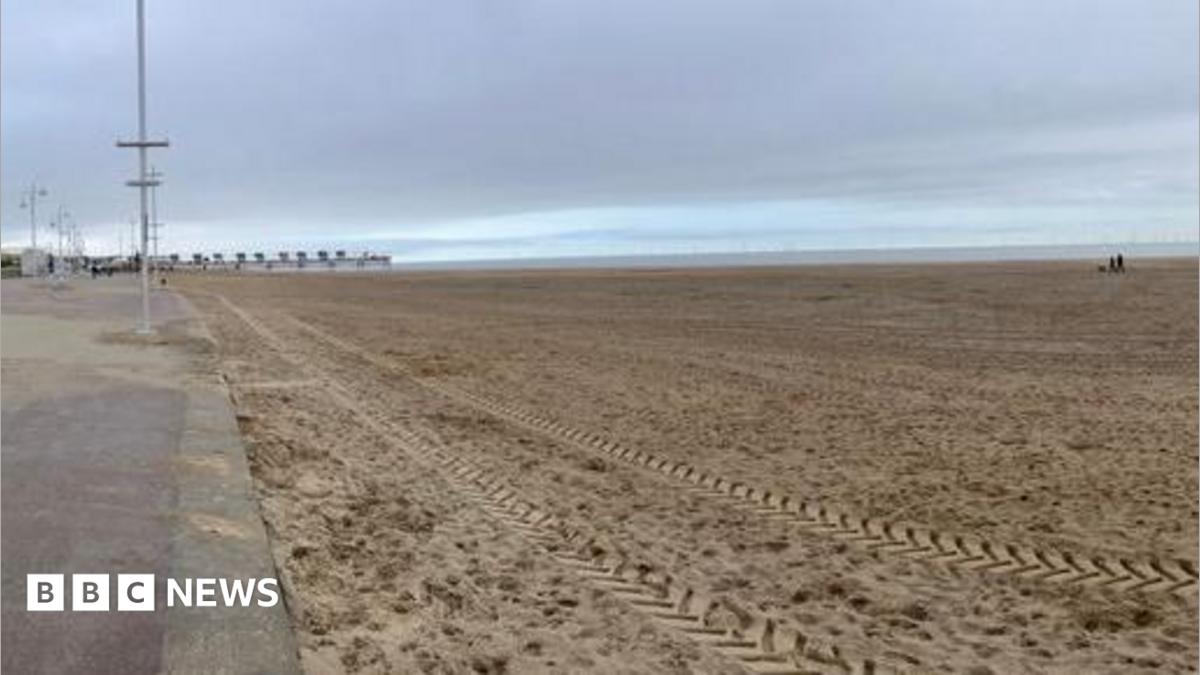A view of the beach on a cloudy day.  A vehicle has left tracks in the sand in the foreground. Part of the promenade is in the left hand side of the picture.  The beach is deserted apart from a couple walking their dog in the distance.