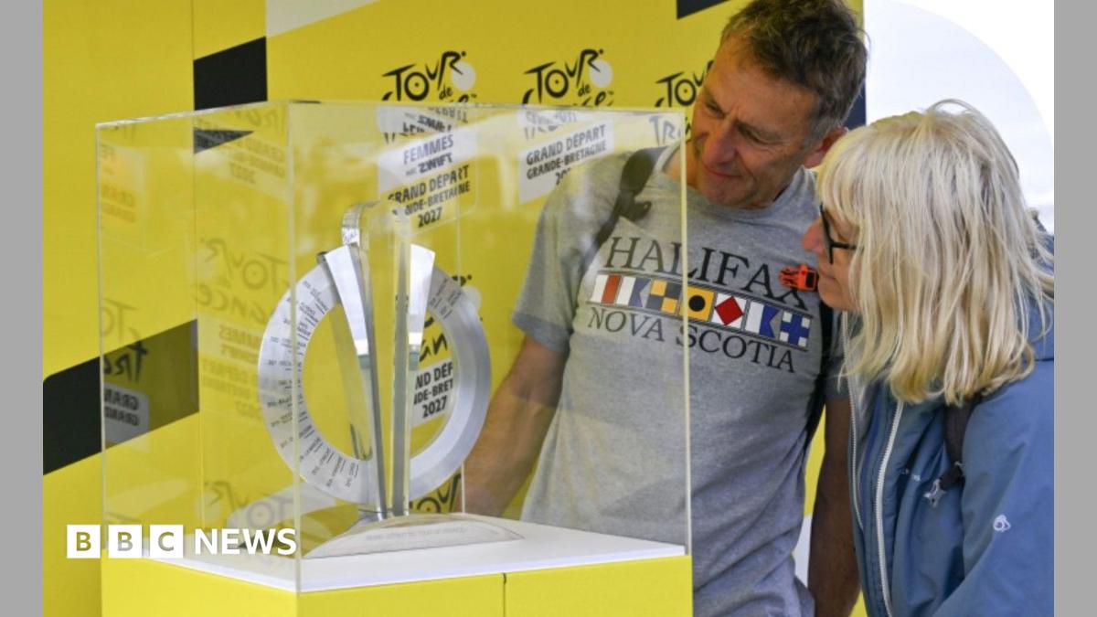 A woman with shoulder length blond hair and glasses, wearing a blue jacket and a man with short dark hair wearing a grey t-shirt with "Nova Scotia" written on it  are both looking at the trophy which is on a yellow plinth and covered with a clear glass or plastic case.