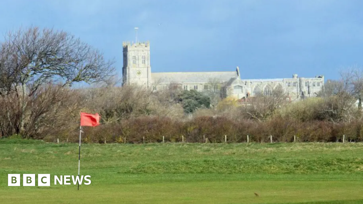 A telephoto shot of Christchurch Priory in the distance, with a putting green and an orange flag of Solent Meads gold course in the foreground. The priory church is a huge stone medieval building with a square tower at one end.